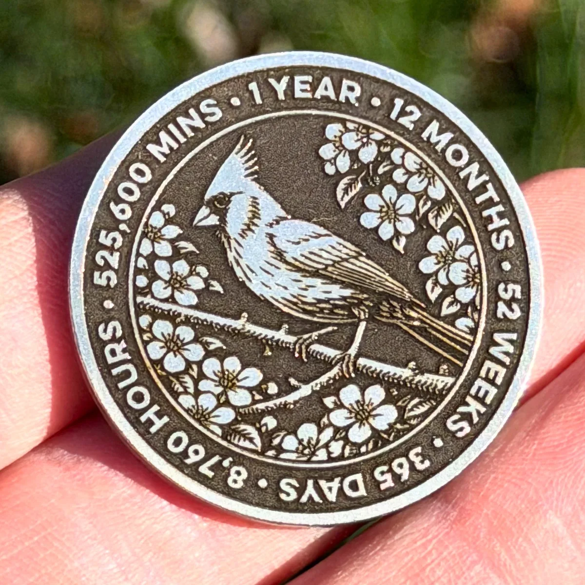 Photo of a sobriety coin held in a hand showing a cardinal bird surrounded by flowers and text marking time milestones