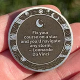 Photo of a silver and dark metal sobriety coin held in a hand, featuring a crescent moon and stars design with a Leonardo Da Vinci quote about fixing your co...